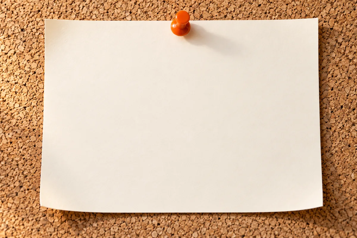 A single sheet of paper pinned to a cork board with one orange pin, in soft side light